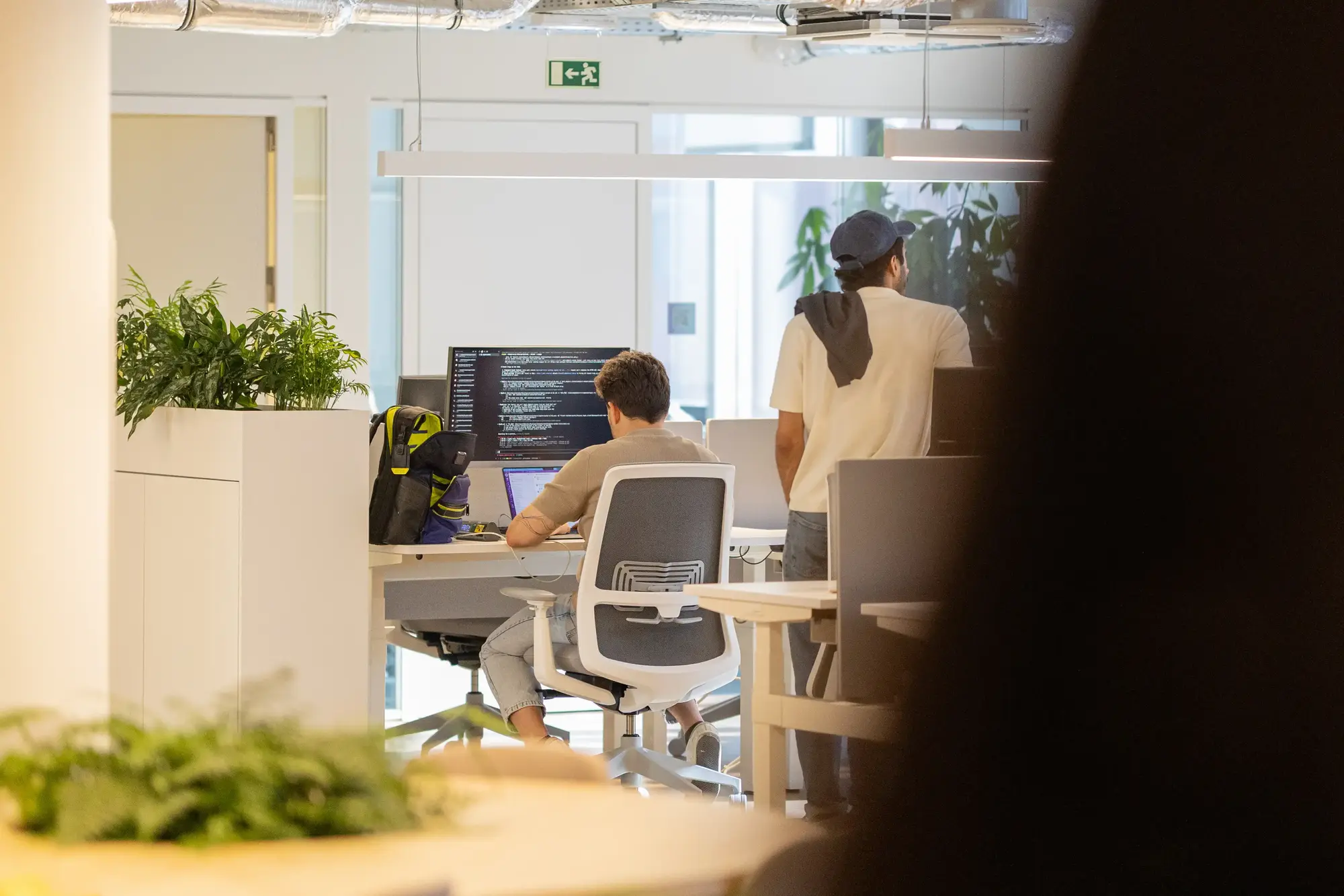 Person walking close by to camera, partially obscuring busy office filled with computer stations.