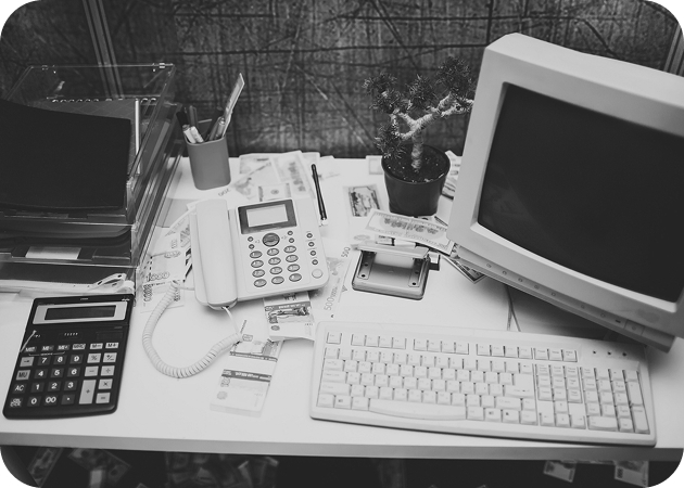 Cluttered desk with phone, calculator, and old CRT computer; black and white