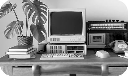 A desk covered in old equipment - computer, cassette player, phone. Black and white