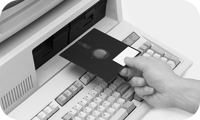 Person inserting a floppy disk drive into an older computer; black and white